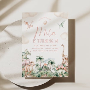 May include: A pink and white dinosaur-themed birthday invitation for a girl turning three. The invitation features a watercolor illustration of dinosaurs, a volcano, and palm trees. The text reads "Stomp on Over Mila is turning 3!" with the date, time, and location of the party.