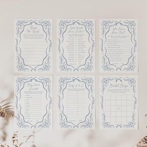 Pu&ograve; includere: Sei biglietti per giochi di baby shower con bordi floreali blu e testo. Le carte includono giochi come "Indovina l'ospite" e "Bingo nuziale". Le carte sono disposte su una superficie bianca con piante secche in primo piano.