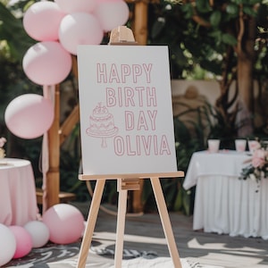 以下が含まれることがあります： 木製のイーゼルには、ピンク色の文字で「HAPPY BIRTH DAY OLIVIA」とケーキのイラストが描かれた白い看板が飾られています。ピンク色の風船とピンク色のテーブルクロスが敷かれたテーブルがあり、誕生日のテーマを示唆しています。