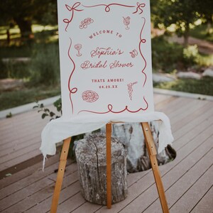 May include: A white sign on a wooden easel with red decorative elements and text. The sign reads "Welcome to Sophia's Bridal Shower" with the date 04.25.XX. The sign is decorated with illustrations of cocktails, pasta, and other food items.