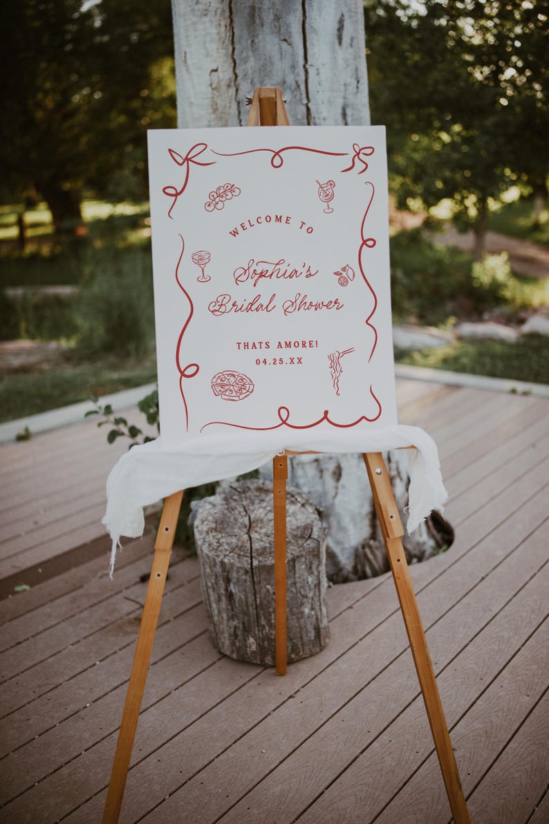 May include: A white sign on a wooden easel with red decorative elements and text. The sign reads "Welcome to Sophia's Bridal Shower" with the date 04.25.XX. The sign is decorated with illustrations of cocktails, pasta, and other food items.
