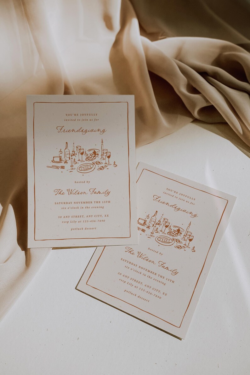 K&ouml;nnte beinhalten: Zwei cremefarbene Friendsgiving-Einladungskarten mit braunem Rand und Text. Die Einladungen enthalten den Text "You're joyfully invited to join us for Friendsgiving" und "The Wilson Family". Die Einladungen liegen auf einer beigen Oberfl&auml;che.
