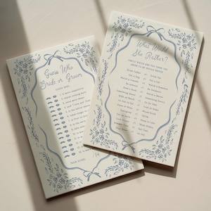 May include: Two rectangular paper game cards with cream backgrounds and blue ink. One card is titled Guess Who Bride or Groom, the other What Would She Rather. Both feature decorative blue floral borders, ribbon accents, and lists of questions.