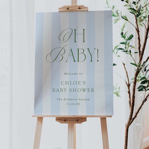 Pu&ograve; includere: Un cavalletto in legno espone un cartello a righe verticali blu e bianche. Il cartello recita "OH BABY!" in elegante corsivo, seguito da "Welcome to CHLOE'S BABY SHOWER" e dettagli dell'evento. Una pianta decorativa &egrave; sullo sfondo.