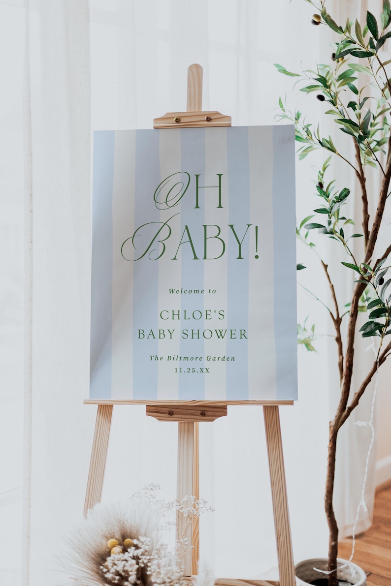 Pu&ograve; includere: Un cavalletto in legno espone un cartello a righe verticali blu e bianche. Il cartello recita "OH BABY!" in elegante corsivo, seguito da "Welcome to CHLOE'S BABY SHOWER" e dettagli dell'evento. Una pianta decorativa &egrave; sullo sfondo.