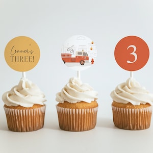 May include: Three cupcakes with white frosting and decorative toppers. One topper says "Conner's THREE!" in black script on a gold circle. Another features a red fire truck illustration. The third has the number "3" on an orange circle.