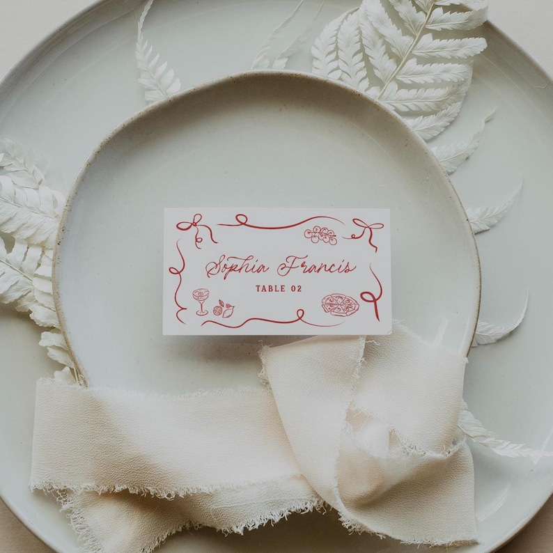 May include: A white place setting with a decorative name card. The card features the name "Sophia Francis" and "TABLE 02" in a burgundy script, along with decorative illustrations. A cream-colored ribbon and white fern are also visible.