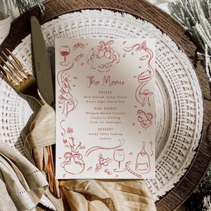 Puede incluir: Una tarjeta de menú festiva con ilustraciones y texto en rojo, que incluye una copa de vino, un árbol de Navidad y un hombre de jengibre. El menú enumera entrantes, guarniciones y postres. La tarjeta está sobre un plato blanco con un tenedor y un cuchillo dorados.