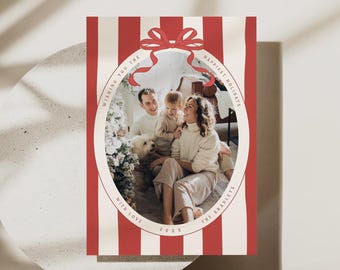 May include: A rectangular holiday greeting card featuring a red and white vertical striped background. An oval frame with a red bow at the top contains a photograph of a family with a dog. The text reads WISHING YOU THE HAPPIEST HOLIDAYS WITH LOVE THE BRADLEYS 20XX.