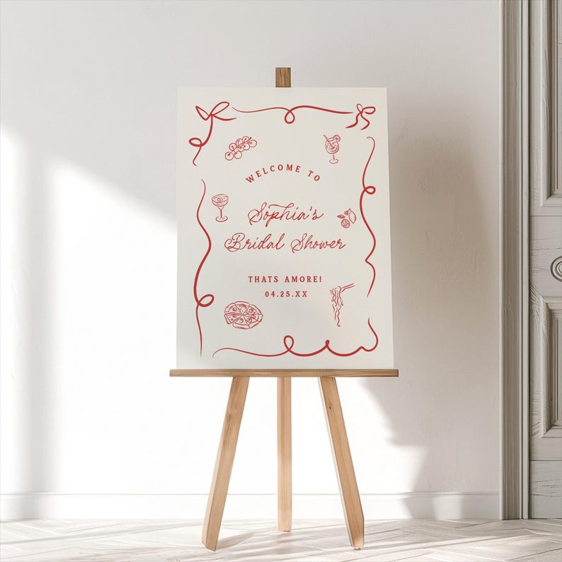 May include: A cream-colored sign on a wooden easel with red decorative elements and text. The sign reads "Welcome to Sophia's Bridal Shower" with the phrase "Thats Amore!" and a date. The design includes food and drink illustrations.