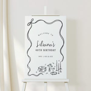 May include: A white sign on an easel with black text and illustrations. The sign reads "Welcome to Liliana's 40th Birthday" with the location and date below. The sign is decorated with a black ribbon border and illustrations of a cake, flowers, and candles.