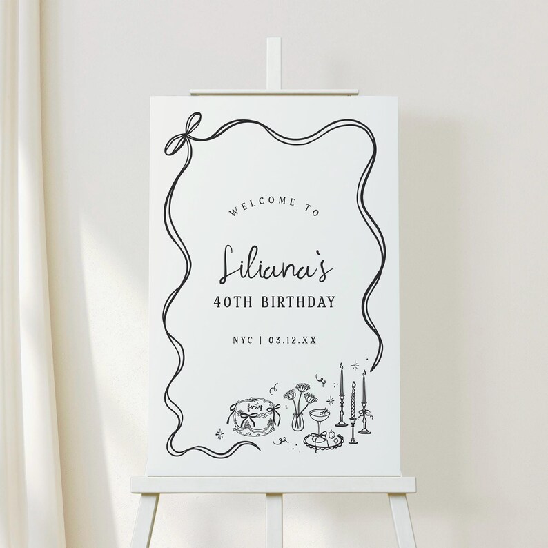 May include: A white sign on an easel with black text and illustrations. The sign reads "Welcome to Liliana's 40th Birthday" with the location and date below. The sign is decorated with a black ribbon border and illustrations of a cake, flowers, and candles.