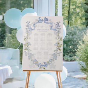 May include: A white rectangular seating chart sign on a wooden easel, featuring a blue watercolor floral wreath and ribbon design. The text reads Sophia's BABY SHOWER and lists tables one through eight with placeholders for guest names.