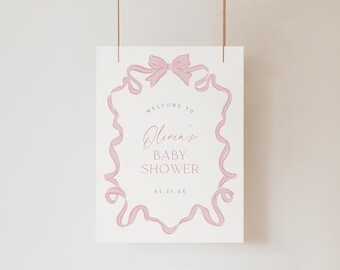 May include: A rectangular white sign featuring a pink ribbon border that forms a frame with a bow at the top. The text reads: WELCOME TO Olivia's BABY SHOWER 04.25.XX. The sign is suspended by two thin copper-colored wires from the top edge.