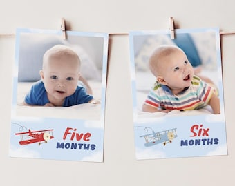 May include: Two rectangular photo prints hanging on a string. The left print shows a baby in a blue shirt with a red airplane graphic and the text Five MONTHS. The right print shows a baby in a striped shirt with a blue airplane graphic and the text Six MONTHS.