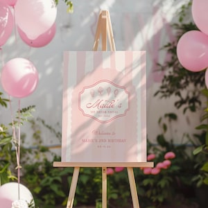 Puede incluir: Un cartel rosa y blanco con un logotipo rosa de cono de helado que dice "Maddie's Ice Cream Social" y "Bienvenido al 2&ordm; cumplea&ntilde;os de Maddie". El cartel est&aacute; en un caballete de madera frente a un arbusto verde y globos rosas.
