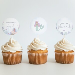 Puede incluir: Tres cupcakes con glaseado blanco cubiertos con adornos redondos de papel para cupcakes. Los adornos muestran una ilustración de una sirena, el texto "Let's be Mermaids" y el texto "Madeline's five!"