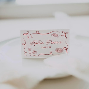 May include: A white place card with red decorative elements and text. The card features the name "Sophia Francis" and "TABLE 02" in a cursive font. Decorative illustrations of a cocktail, fruit, and pizza are also present.