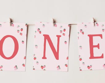 May include: Three rectangular cards hanging from a string with wooden clips. Each card features a light pink background with a pattern of small red strawberries and a large, bold, red letter spelling out the word ONE.