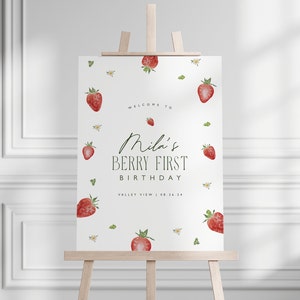 May include: A white sign on a wooden easel, decorated with watercolor strawberries and floral accents. The sign reads "Welcome to Mila's Berry First Birthday" with the date 08.26.24. A charming design for a birthday celebration.