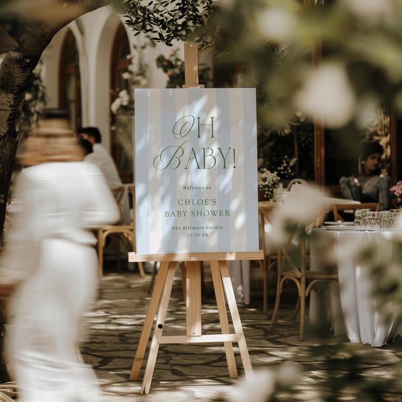 Pu&ograve; includere: Un cavalletto in legno espone un cartello con le parole "OH BABY!" in elegante scrittura. Il cartello ha uno sfondo a righe azzurre e bianche e d&agrave; il benvenuto agli invitati del baby shower di Chloe.