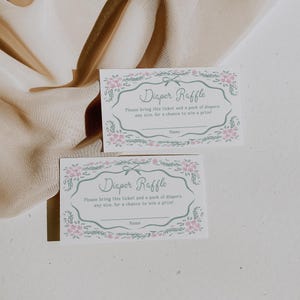 Op de afbeelding: Twee rechthoekige luierlottickets met bloemenranden en de tekst "Diaper Raffle". De tickets hebben ruimte voor een naam en instructies om luiers mee te nemen voor een kans op een prijs. Zachte, neutrale achtergrond.