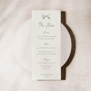 May include: A light-coloured menu card with a bow illustration and the words "The Menu" in script. The menu lists starters, main courses, and dessert. The text "Olivia's Baby Shower" and the date 04.15.23 are at the bottom. The card is on a circular stand.