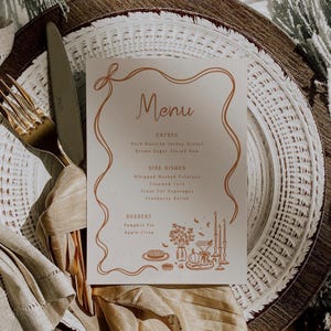 Könnte beinhalten: Eine Menükarte mit dem Wort „Menu“ in eleganter Schrift, die Gerichte wie Kräuterbrust, braun glasierter Schinken und Kartoffelpüree auflistet. Die Karte liegt auf einem weißen Teller mit einer goldenen Gabel und einem Messer.