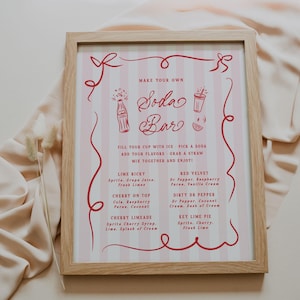 May include: A framed sign with a pink and white striped background and red text that reads "Make Your Own Soda Bar." The sign lists soda flavor combinations like Lime Ricky and Cherry on Top. The frame is made of light-colored wood.