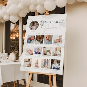 以下が含まれることがあります： 「One Year of Jackson」のパーソナライズされた写真ディスプレイボード。ボードには、赤ちゃんの写真がグリッド状に配置され、熱気球のグラフィックと赤ちゃんの名前がエレガントなスクリプトで表示されています。ボードは木製のイーゼルに表示されています。