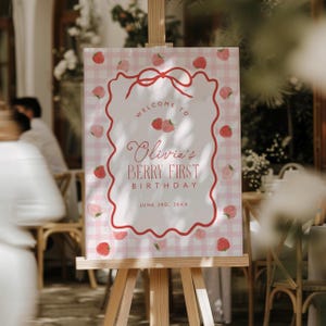 May include: A welcome sign for a first birthday party. The sign features a pink gingham background with a red border and strawberry illustrations. Text reads "Welcome to Olivia's Berry First Birthday" and the date.