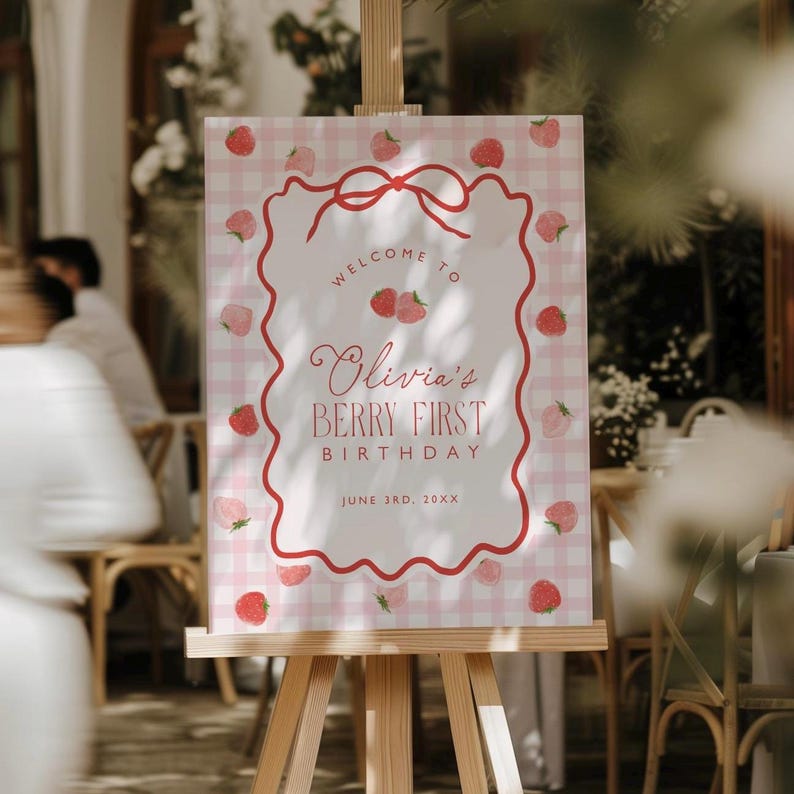 May include: A welcome sign for a first birthday party. The sign features a pink gingham background with a red border and strawberry illustrations. Text reads "Welcome to Olivia's Berry First Birthday" and the date.