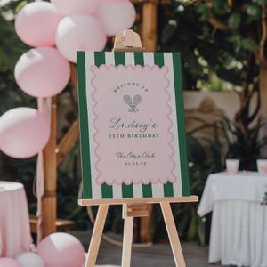 May include: A wooden easel displays a sign with a pink and green tennis theme. The sign reads "Welcome to Lindsey's 25th Birthday" with tennis rackets and balls. Pink balloons are in the background.