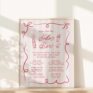 May include: A framed poster with a pink and white striped background and red decorative elements. The poster reads "Make Your Own Soda Bar" and lists soda flavor combinations like Lime Ricky and Red Velvet. The poster is displayed on a light-colored surface.