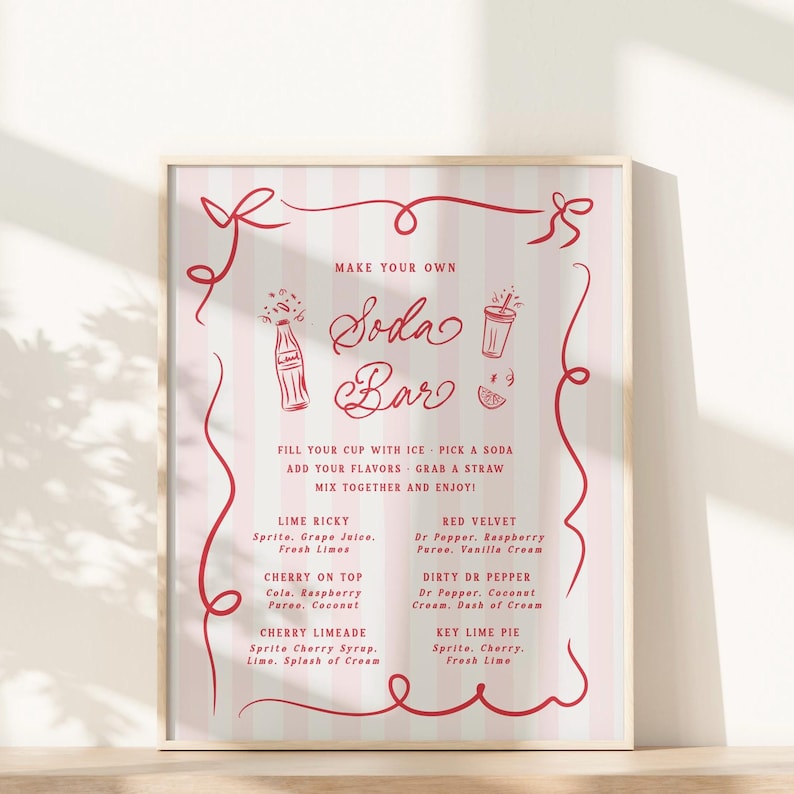 May include: A framed poster with a pink and white striped background and red decorative elements. The poster reads "Make Your Own Soda Bar" and lists soda flavor combinations like Lime Ricky and Red Velvet. The poster is displayed on a light-colored surface.