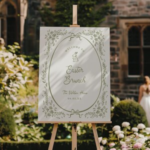 May include: A white sign on a wooden easel, welcoming guests to an Easter Brunch. The sign features an oval frame with floral and bow accents, and the words "Welcome to Easter Brunch" with a bunny illustration. The date is printed at the bottom.