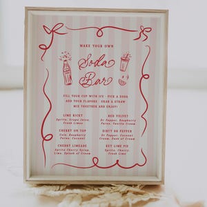 May include: A framed sign with a pink and white striped background and red text. The sign reads "Make Your Own Soda Bar" and lists soda flavor combinations like Lime Ricky and Red Velvet. Decorative red swirls and bows frame the text.