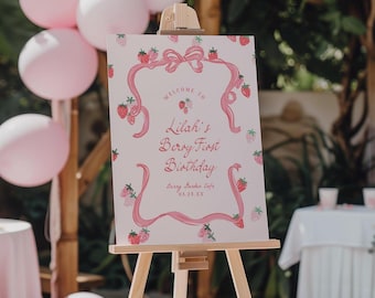 May include: A rectangular sign on a wooden easel featuring a white background with pink strawberry illustrations and a pink ribbon border. The text reads: Welcome to Lilah's Berry First Birthday, Berry Garden Cafe, 03.23.XX.