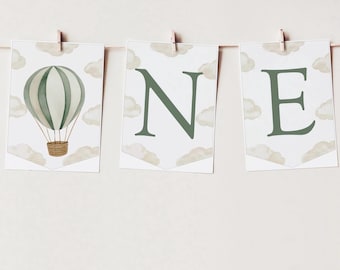 May include: A decorative banner consisting of three white rectangular cards hanging from a string with wooden clips. The cards feature a watercolor hot air balloon and the letters O, N, and E in dark green, surrounded by beige clouds.