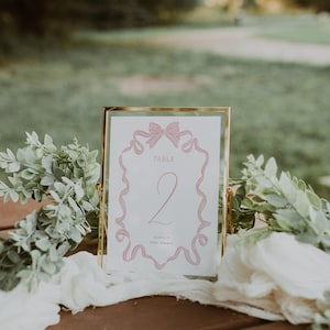 May include: A table number card with a gold frame. The card has a pink ribbon design around the edge and the number 2 in the center. The text "TABLE" and "Olivia's Baby Shower" are also visible. The card is on a wooden surface with greenery.