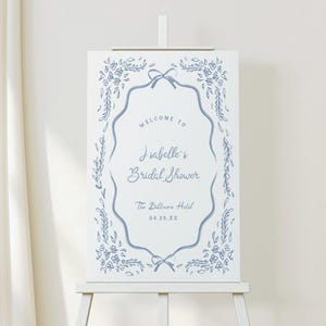 May include: A white welcome sign on an easel with blue floral and bow accents. The sign reads "Welcome to Isabelle's Bridal Shower" and includes the date and location, The Biltmore Hotel.