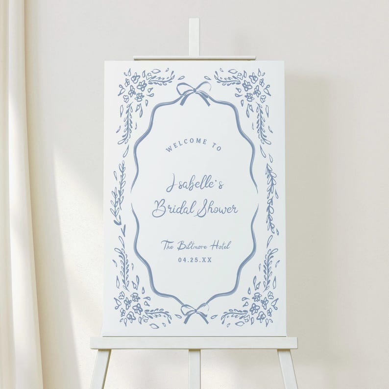 May include: A white welcome sign on an easel with blue floral and bow accents. The sign reads "Welcome to Isabelle's Bridal Shower" and includes the date and location, The Biltmore Hotel.