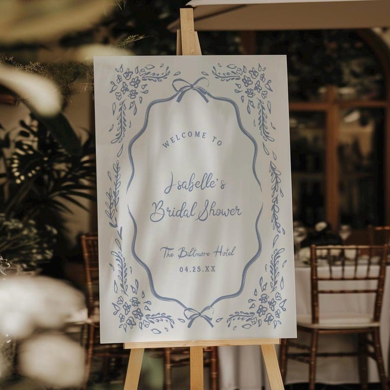 May include: A white sign on an easel with blue floral and ribbon accents. The sign reads "Welcome to Isabelle's Bridal Shower" and includes the date and location, The Biltmore Hotel.