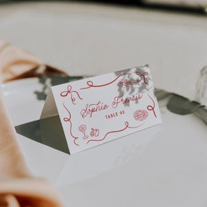 May include: A white place card with red decorative lines and illustrations. The card reads "Sophia Francis" and "TABLE 02" in a cursive font. The card is folded and sits on a white surface.