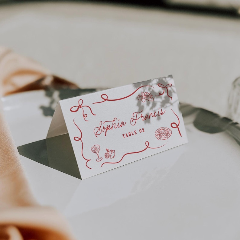 May include: A white place card with red decorative lines and illustrations. The card reads "Sophia Francis" and "TABLE 02" in a cursive font. The card is folded and sits on a white surface.