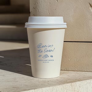 May include: A cream-coloured disposable paper coffee cup with a white plastic lid. The cup features blue text that reads Bun in the Oven!, with small illustrations of baked goods, and the text Sophia's Baby Shower 04.25.XX printed below.