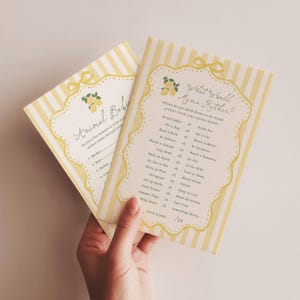 May include: Two rectangular baby shower game cards with yellow and white vertical stripes. The cards feature a scalloped border with a bow at the top and lemon illustrations. The text reads Animal Babies and What Would Mama Rather with game prompts.