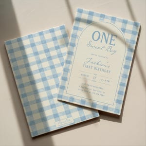 May include: Two light blue and white gingham patterned invitations. The invitation on the right has text that reads "ONE Sweet Boy" and "Jackson's FIRST BIRTHDAY". The date is August 15th, with the time 12-3 PM. The bottom left corner of the invitation on the left has the text "Paper Edit".