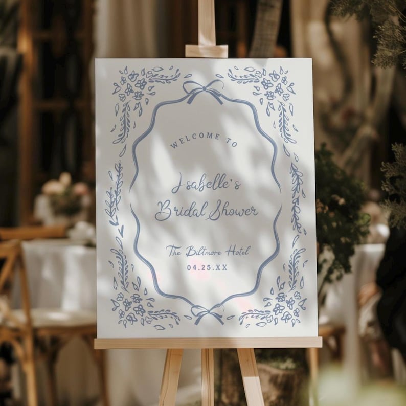 May include: A white sign on a wooden easel with blue floral and leaf border. The sign reads "Welcome to Isabelle's Bridal Shower" and includes the date and location, The Biltmore Hotel.