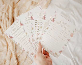May include: A set of white paper baby shower game cards featuring floral illustrations in pink, purple, and green. The cards include text such as Baby Smith Predictions, Baby Animal Match, and Family Feud Baby Edition with blank lines and checkboxes for guests to fill out.